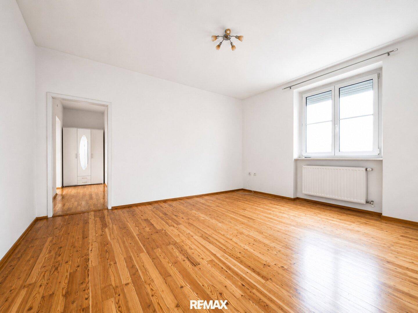 Helles, leeres Zimmer mit Holzboden, weißen Wänden und einem Fenster mit Heizkörper.