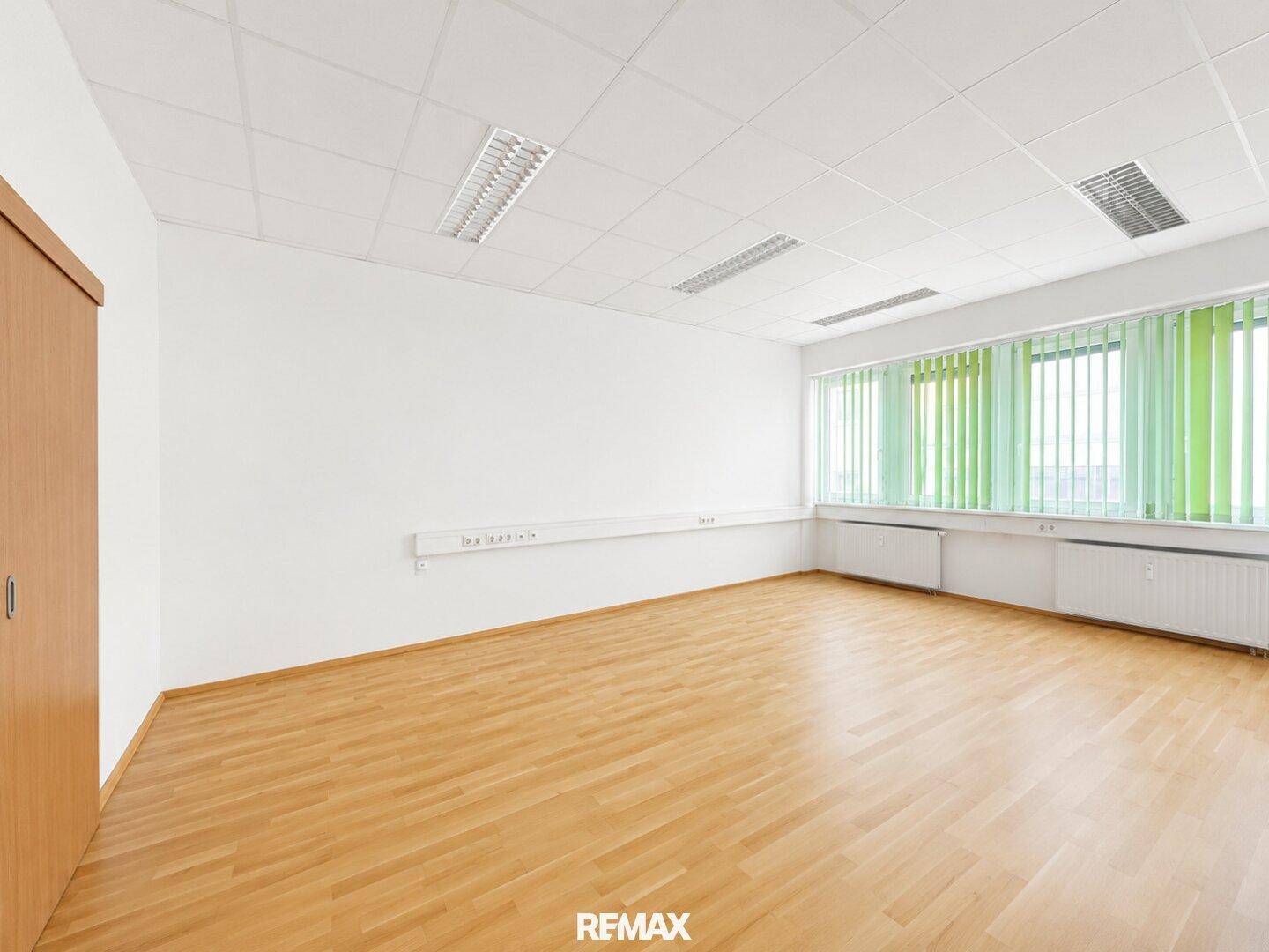 Großes Bürozimmer mit hellem Holzparkett, großen Fenstern und grünen Jalousien.