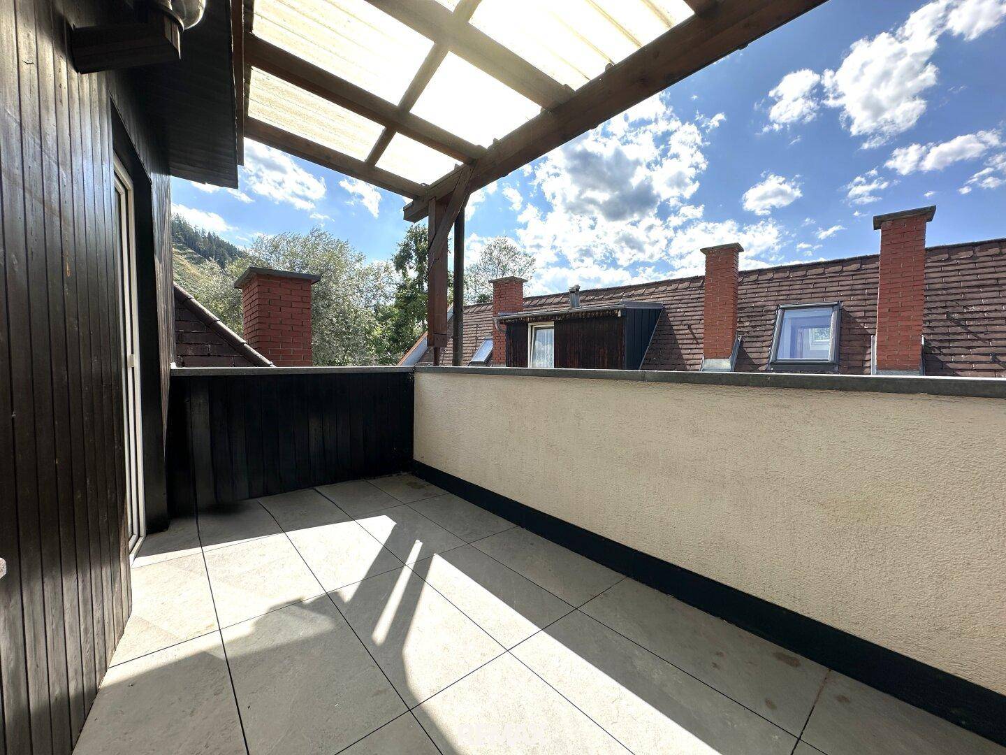 Überdachte Terrasse mit Fliesenboden und Blick auf die umliegenden Dächer und den Himmel.