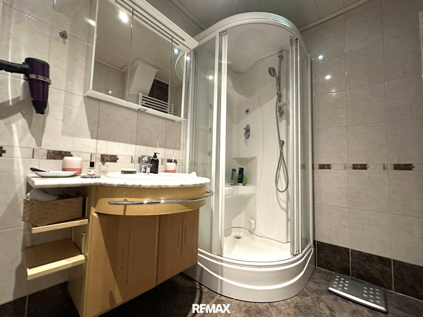 Badezimmer mit Eckduschkabine, Einzelwaschbecken und Spiegelschrank, gefliest in hellen Tönen.