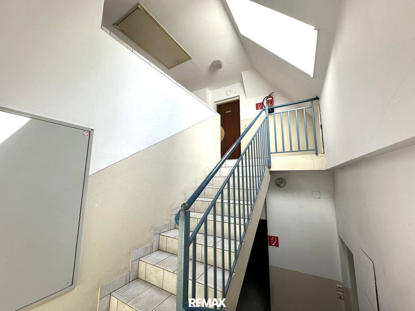 Treppenhaus mit gefliesten Stufen und blauem Metallgeländer, führt zu den oberen Etagen.