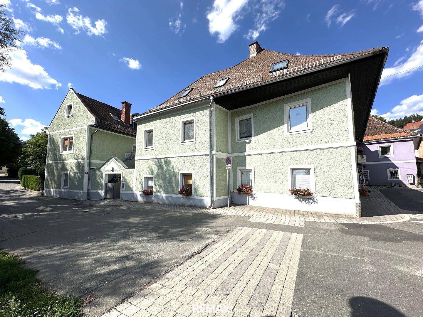 Außenansicht eines hellgrünen Gebäudes mit mehreren Fenstern und einem gepflasterten Vorplatz.