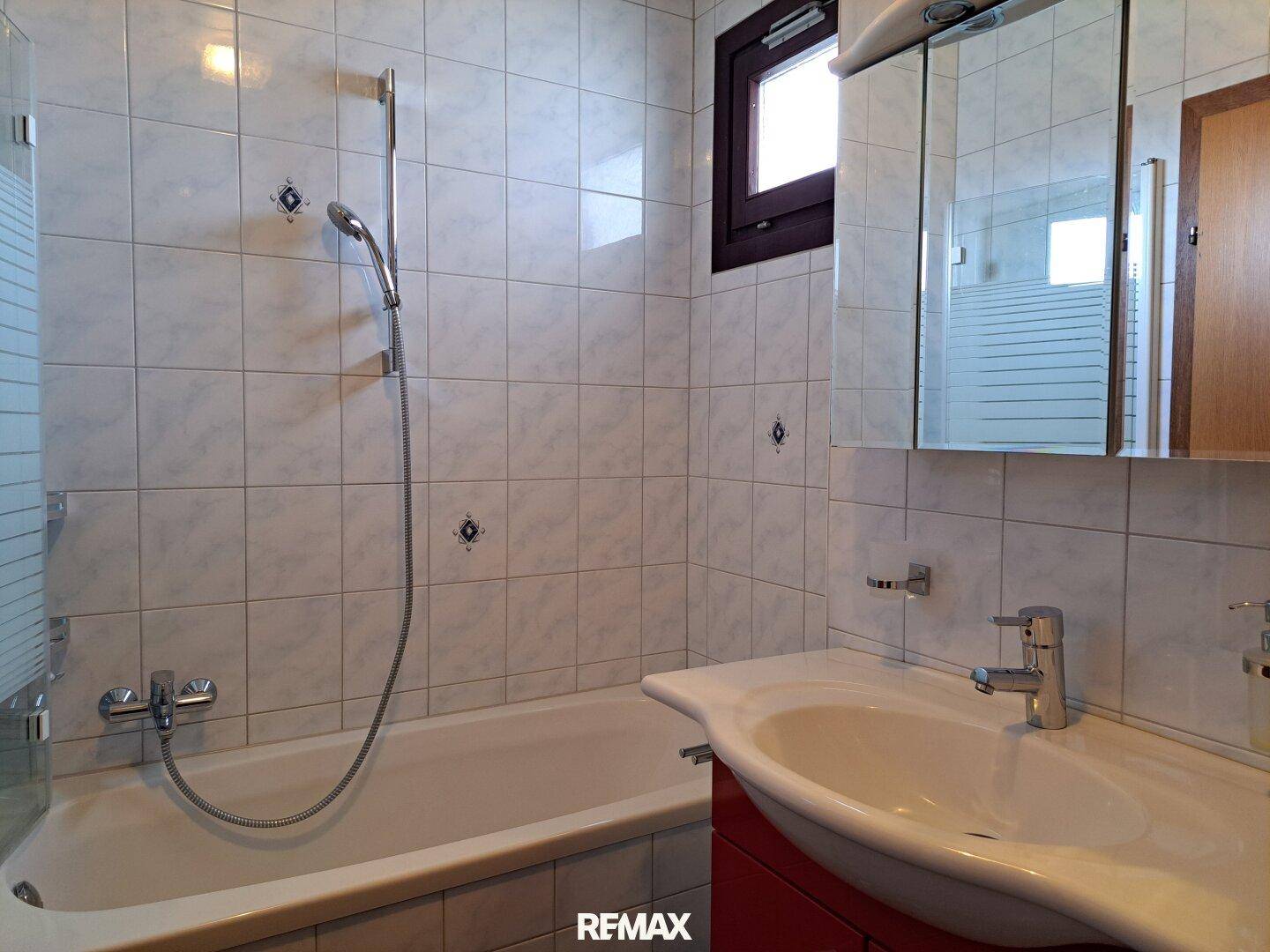 Badezimmer mit Badewanne, Duschvorrichtung, Einzelwaschbecken und Spiegelschrank. Die Fliesen und Armaturen deuten auf einen Stil der 80er Jahre hin.