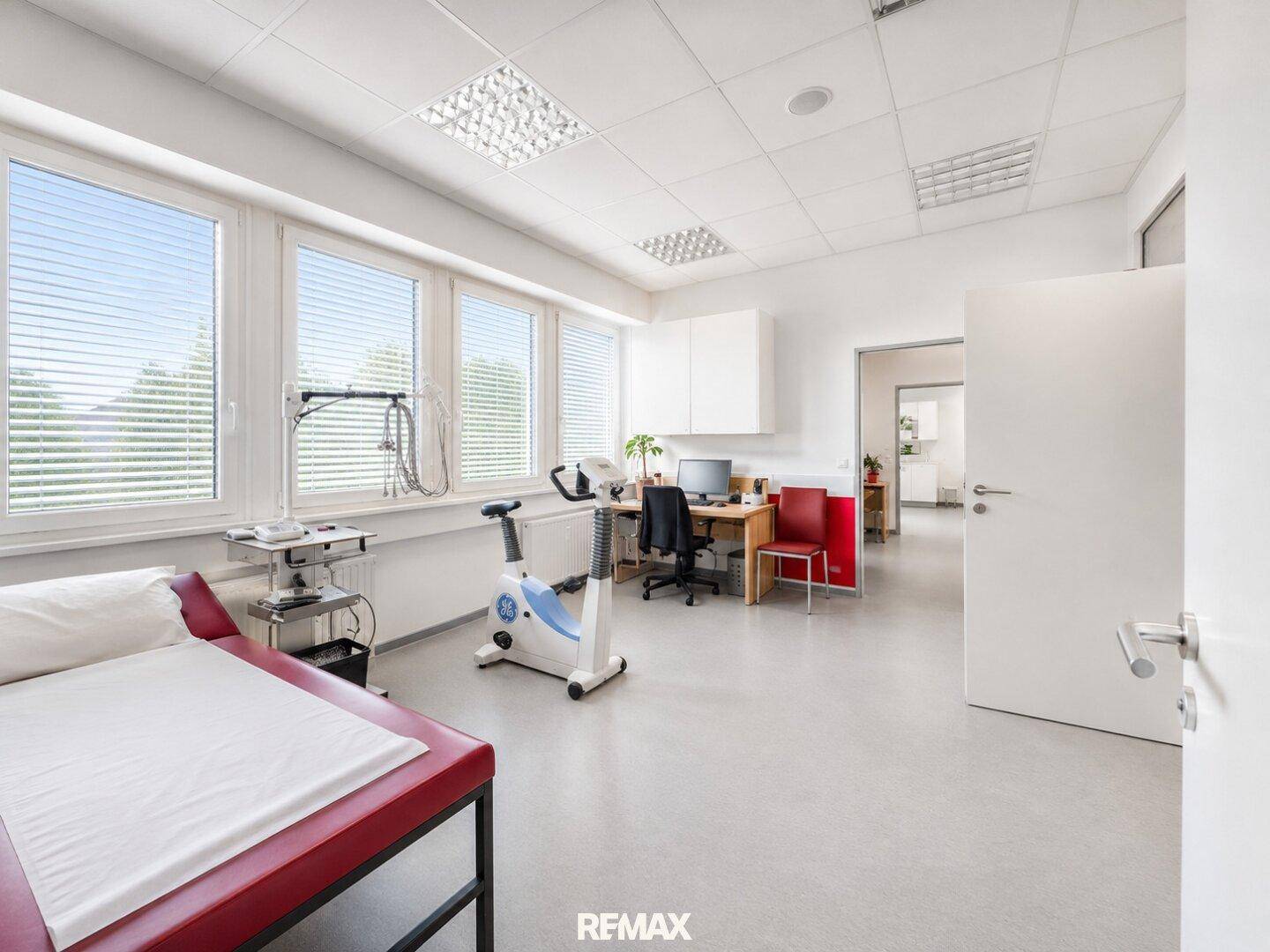 Behandlungsraum mit Untersuchungsliege, Ergometer und Schreibtisch am Fenster.