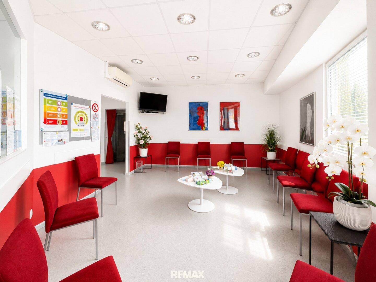 Heller Wartebereich mit roten Stühlen, Couchtischen und Fernseher an der Wand.