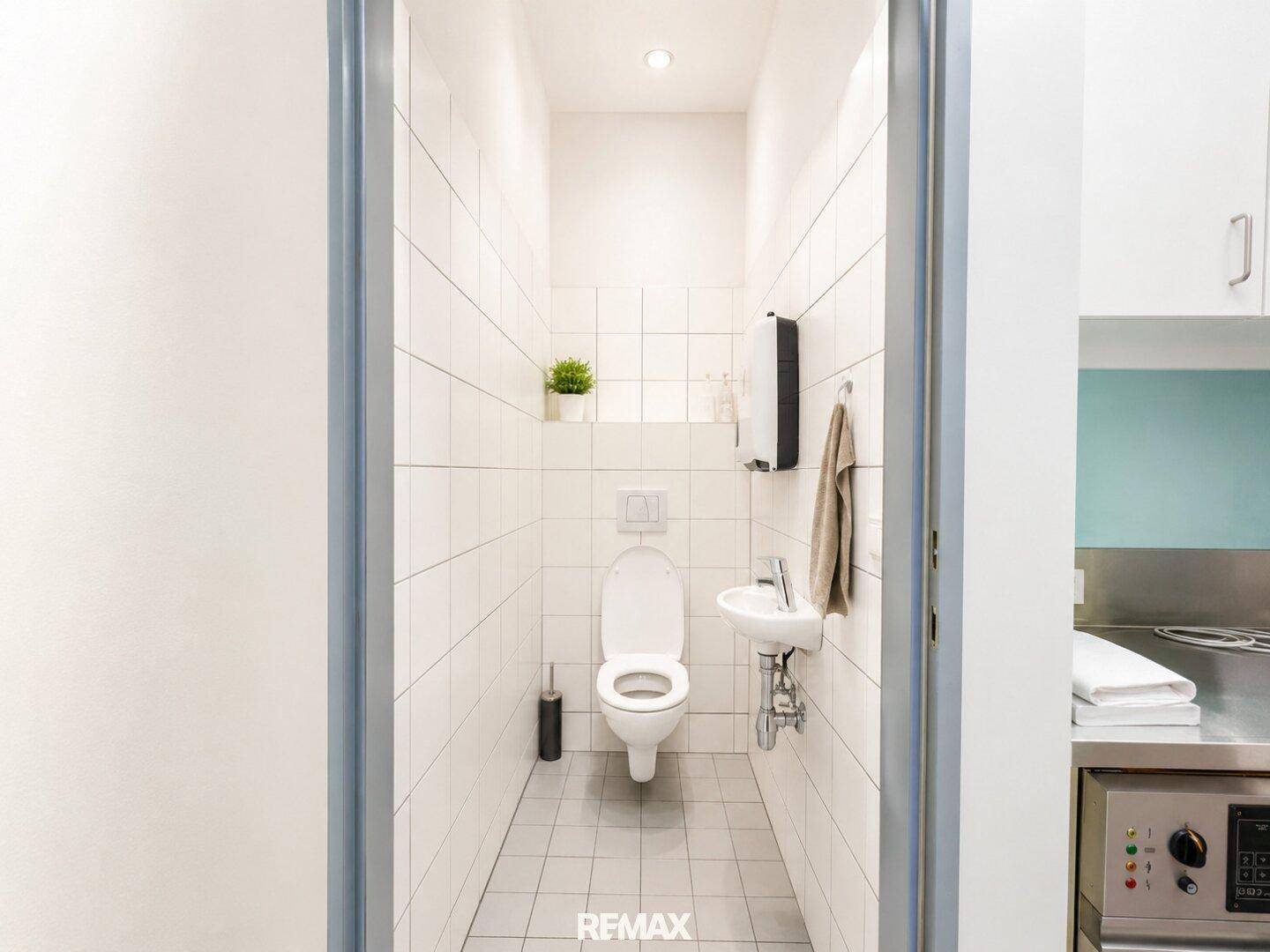 Sauberes WC mit weißen Fliesen, wandmontierter Toilette und kleinem Handwaschbecken.