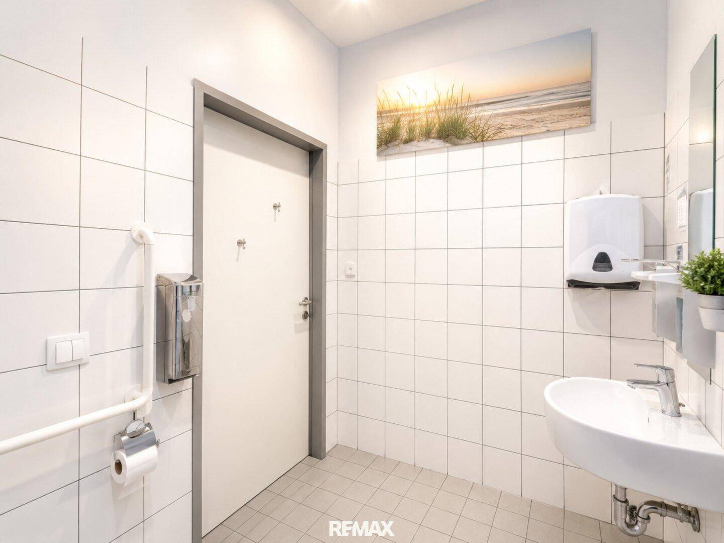 Modernes WC mit weißen Fliesen, Waschbecken und Haltegriffen für Barrierefreiheit.