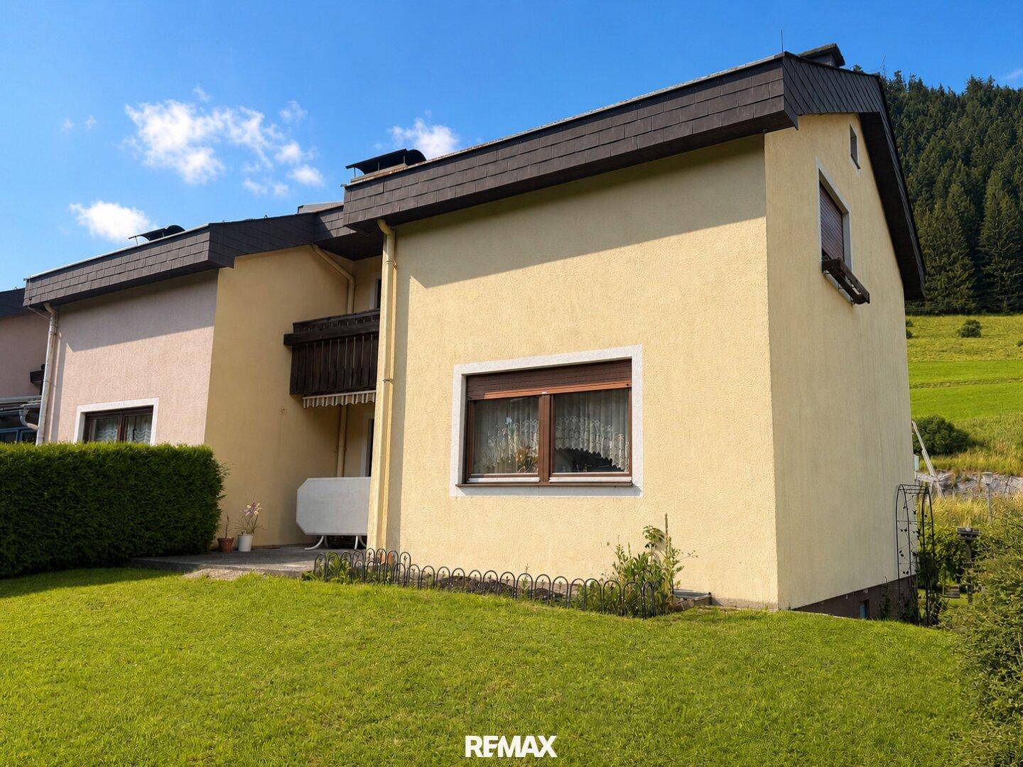 Seitenansicht des Hauses mit gelber Fassade, grünem Rasen und bewaldetem Hintergrund.