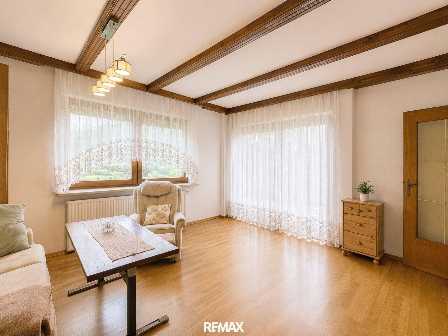 Gemütliches Wohnzimmer mit Holzbalkendecke, Parkettboden und großen Fenstern für viel Licht.
