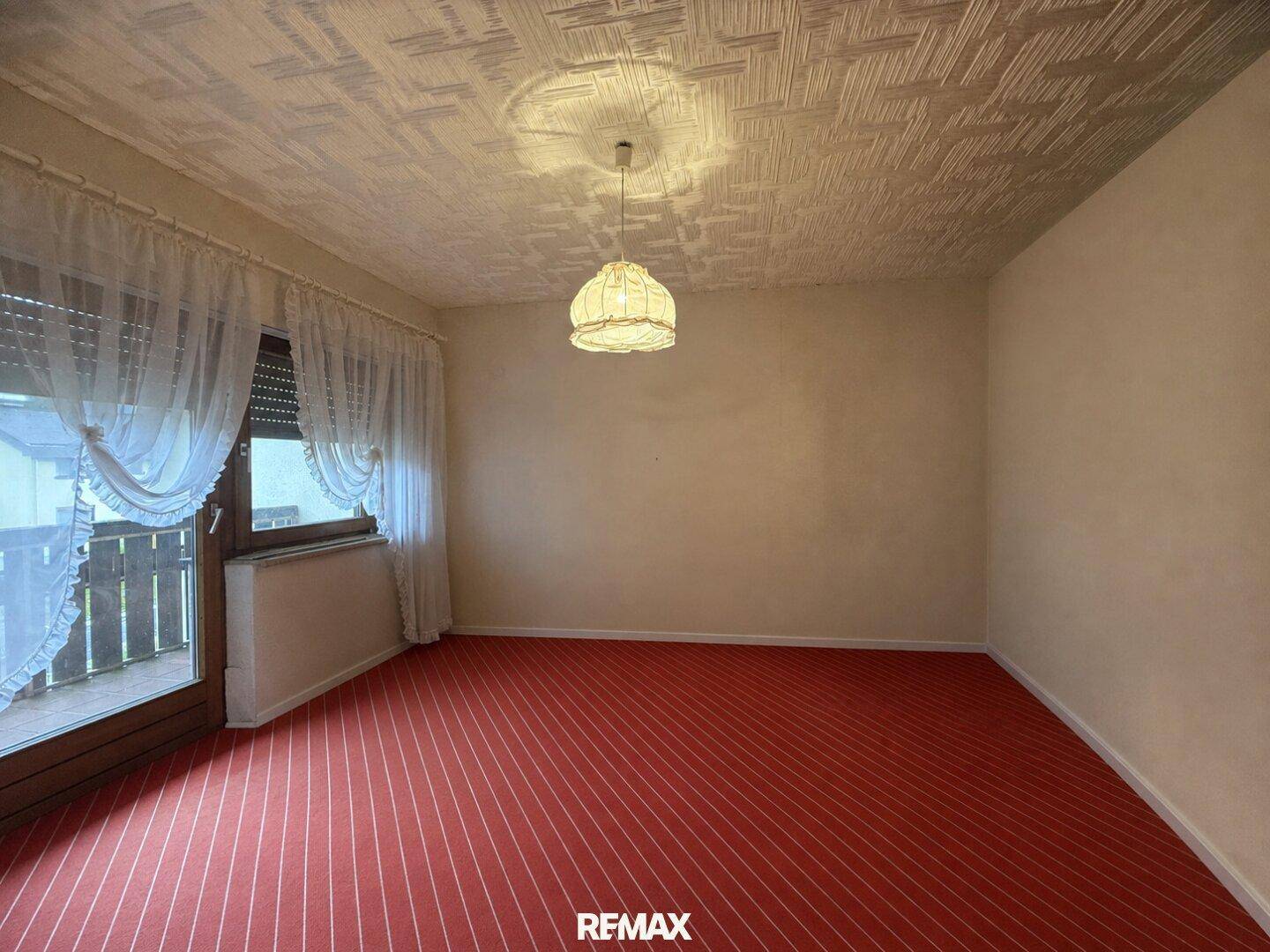 Schlafzimmer mit rotem Teppichboden, strukturiertem Deckenpaneel und Zugang zum Balkon.