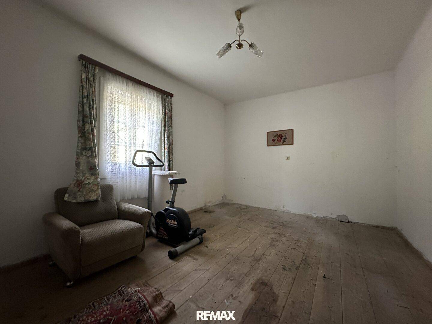 Zimmer mit Holzboden, Fenster und einem Heimtrainer, renovierungsbedürftig.