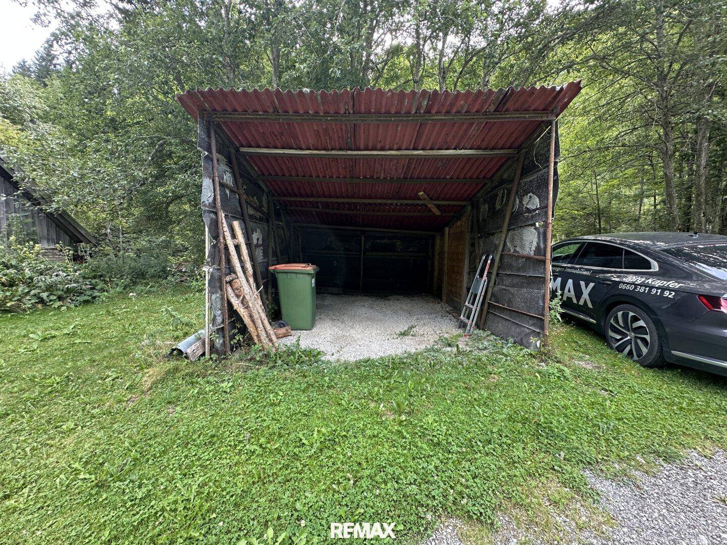 Einfacher Unterstand oder Schuppen im Gartenbereich, umgeben von Bäumen.