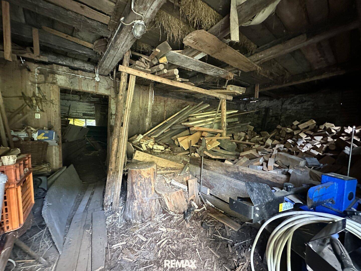 Rustikaler Innenraum mit Holzbalken, Heu und gestapeltem Brennholz, möglicherweise ein Lagerraum oder Stall.