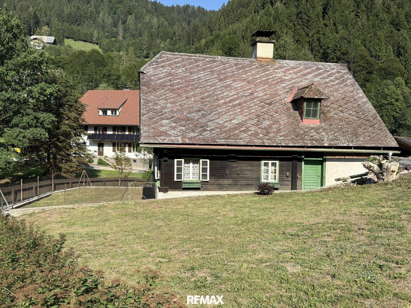 Rustikales Holzhaus mit traditionellem Dach und grüner Wiese im Vordergrund, umgeben von Natur.