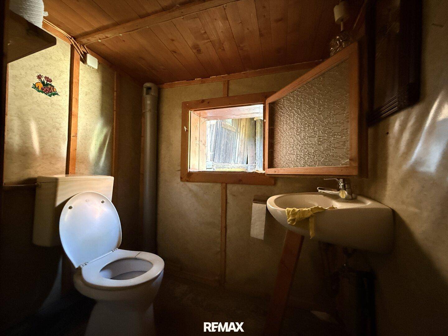 Einfaches WC mit Holzvertäfelung, kleinem Waschbecken und Fenster, funktionaler Raum.