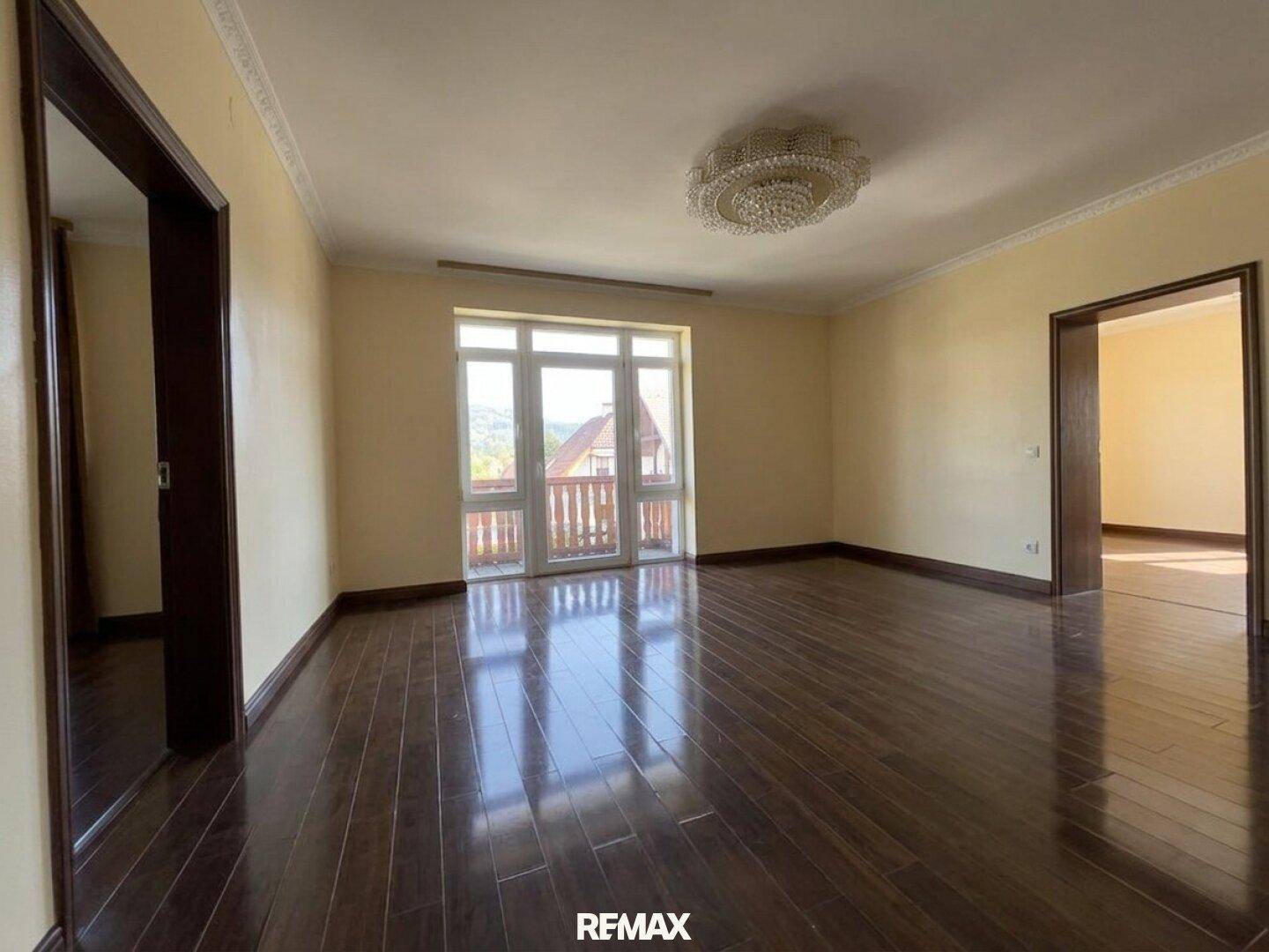 Geräumiger, leerer Raum mit gelben Wänden, dunklem Holzboden und großen Fenstern zum Balkon.