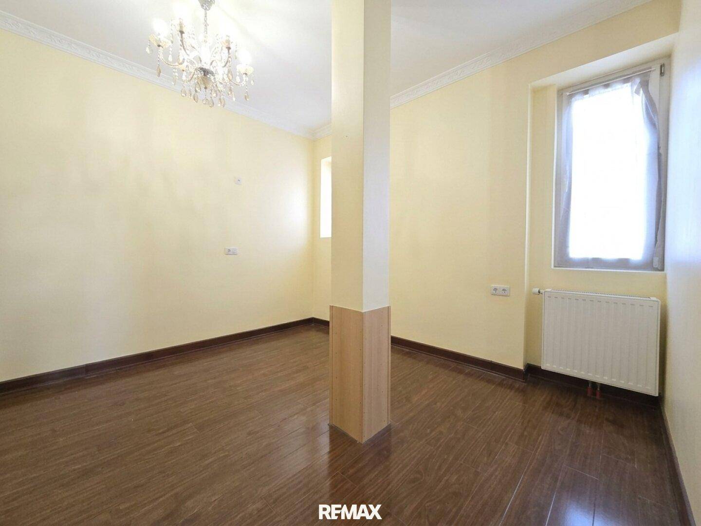 Heller Raum mit gelben Wänden, dunklem Holzboden, einem zentralen Pfeiler und einem Kronleuchter.
