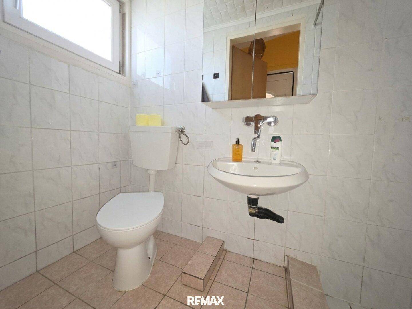 Einfaches WC mit Toilette, Waschbecken und Spiegel, hell gefliest.