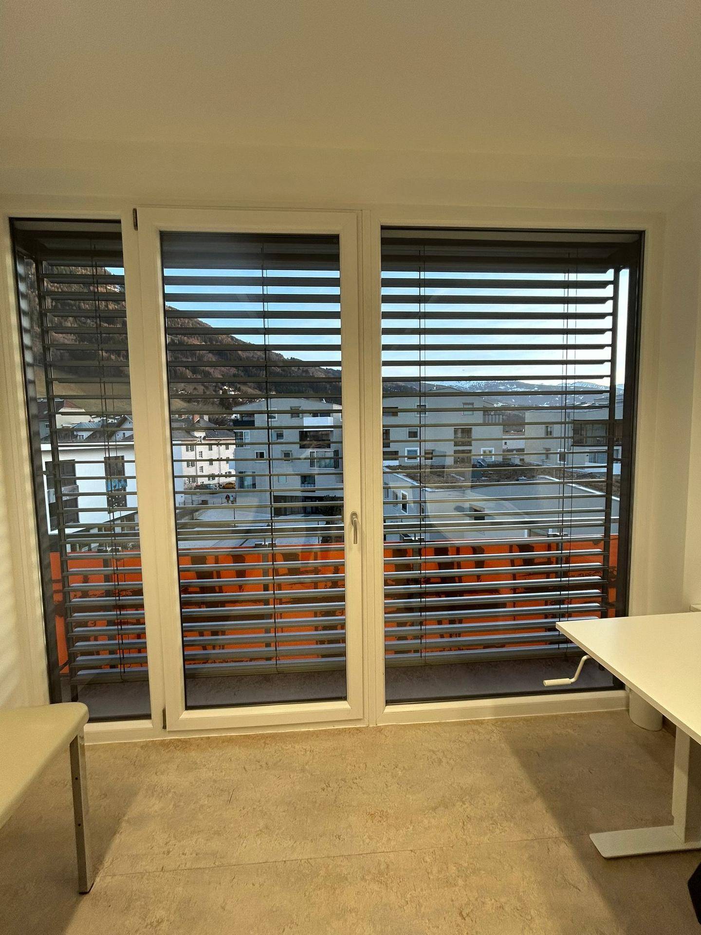 Großes Fenster mit Jalousien bietet einen weiten Blick auf die umliegende Stadtlandschaft und die Berge.