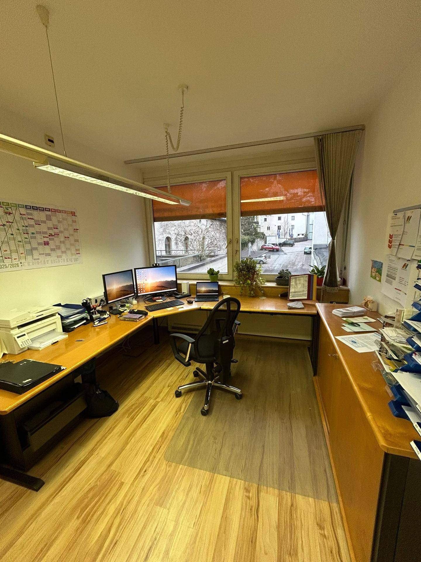 Helles Büro mit L-förmigem Schreibtisch, Holzfußboden und großem Fenster mit Straßenblick.