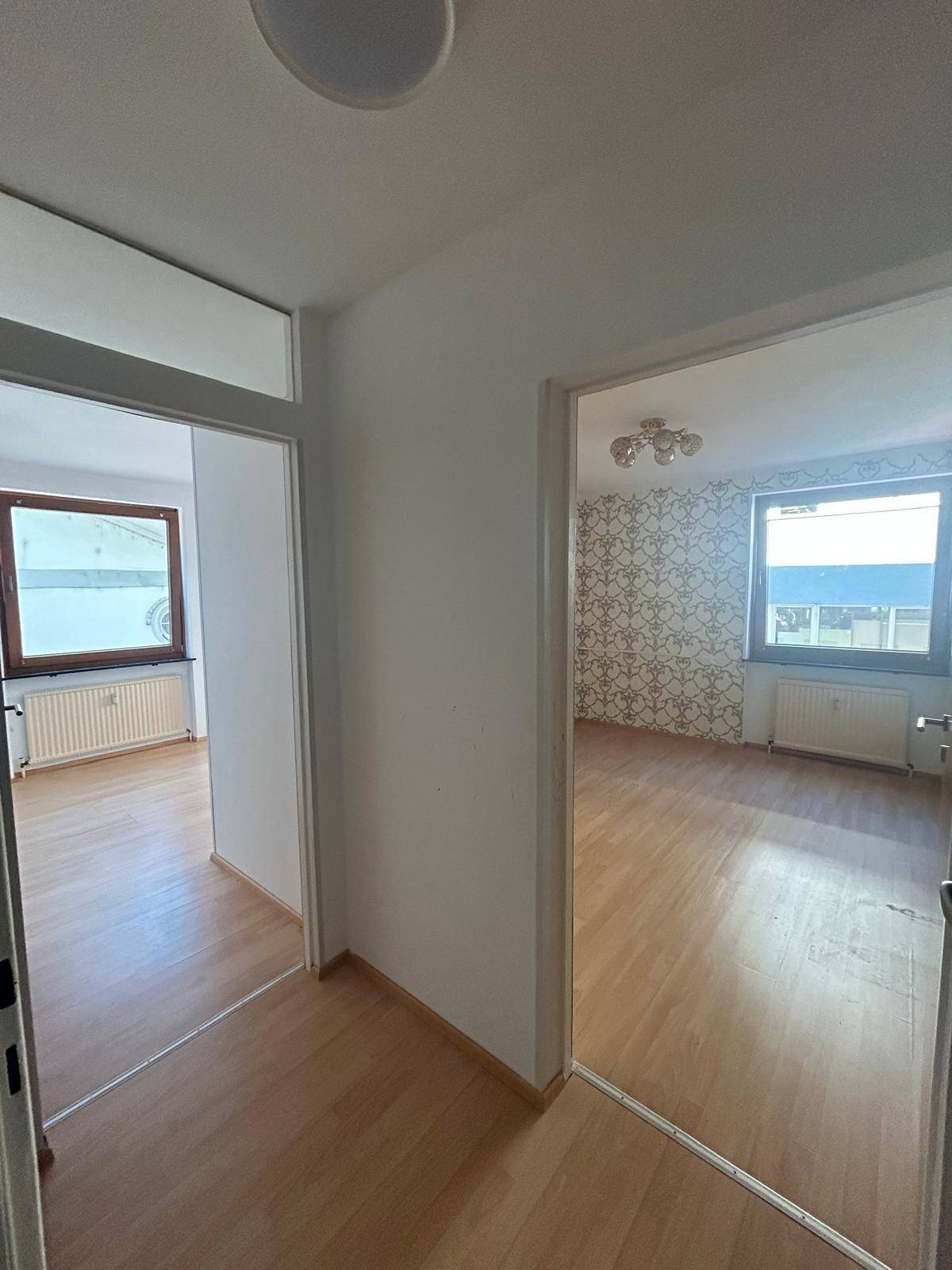Flur mit Laminatboden und Zugang zu zwei Zimmern, eines davon mit gemusterter Tapete und Fenster.
