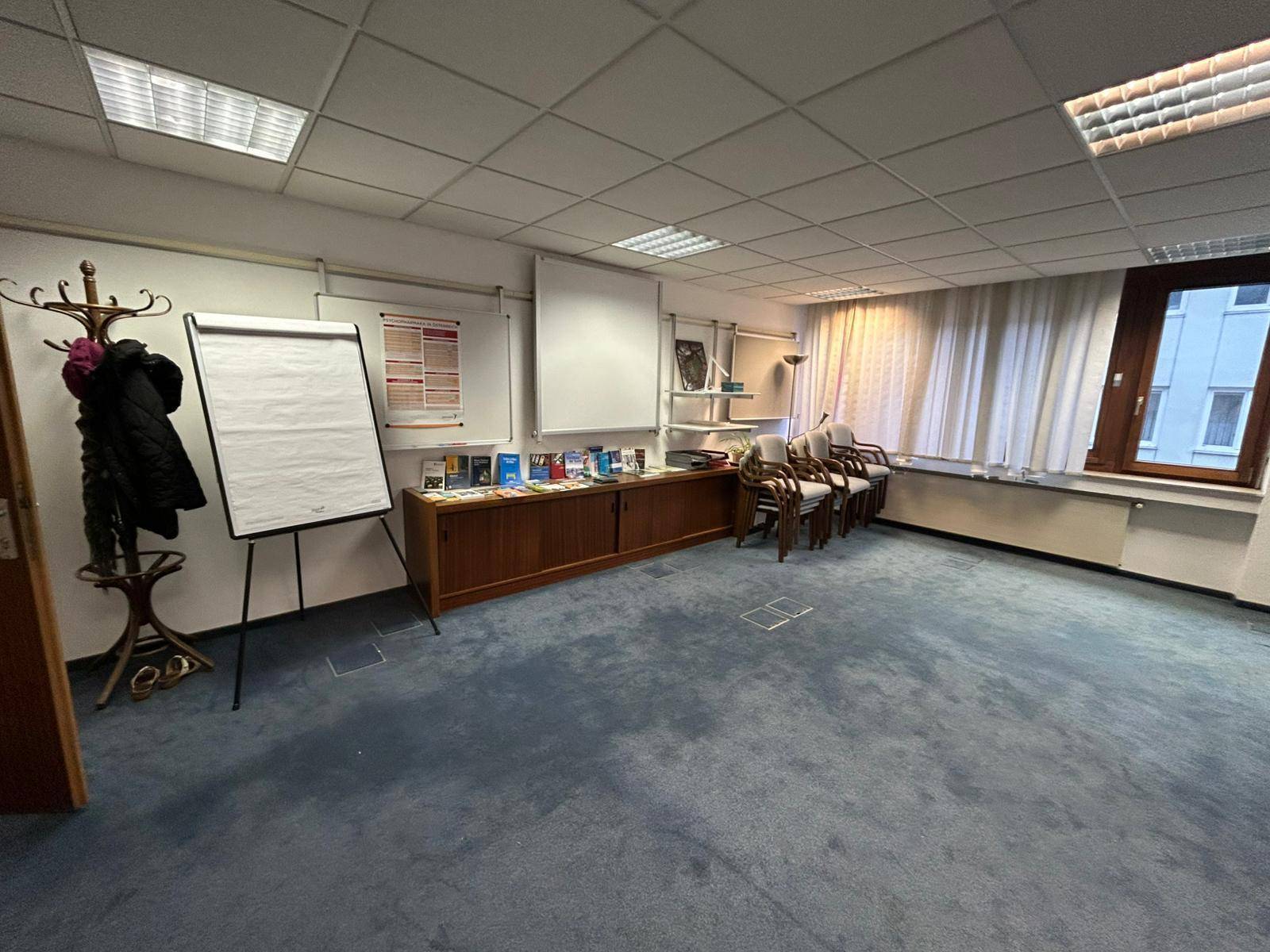 Konferenzraum mit blauem Teppichboden, Flipchart, Schränken und Stühlen für Besprechungen und Präsentationen.