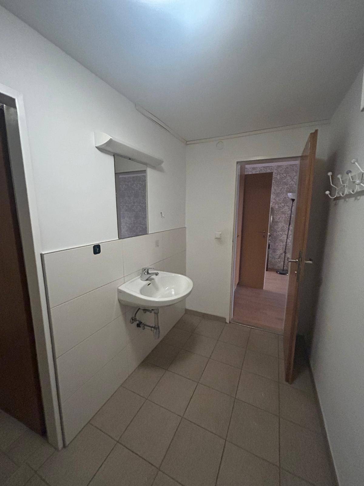 Eingangsbereich mit Waschbecken, Spiegel und Fliesenboden, führt zu weiteren Räumen der Wohnung.