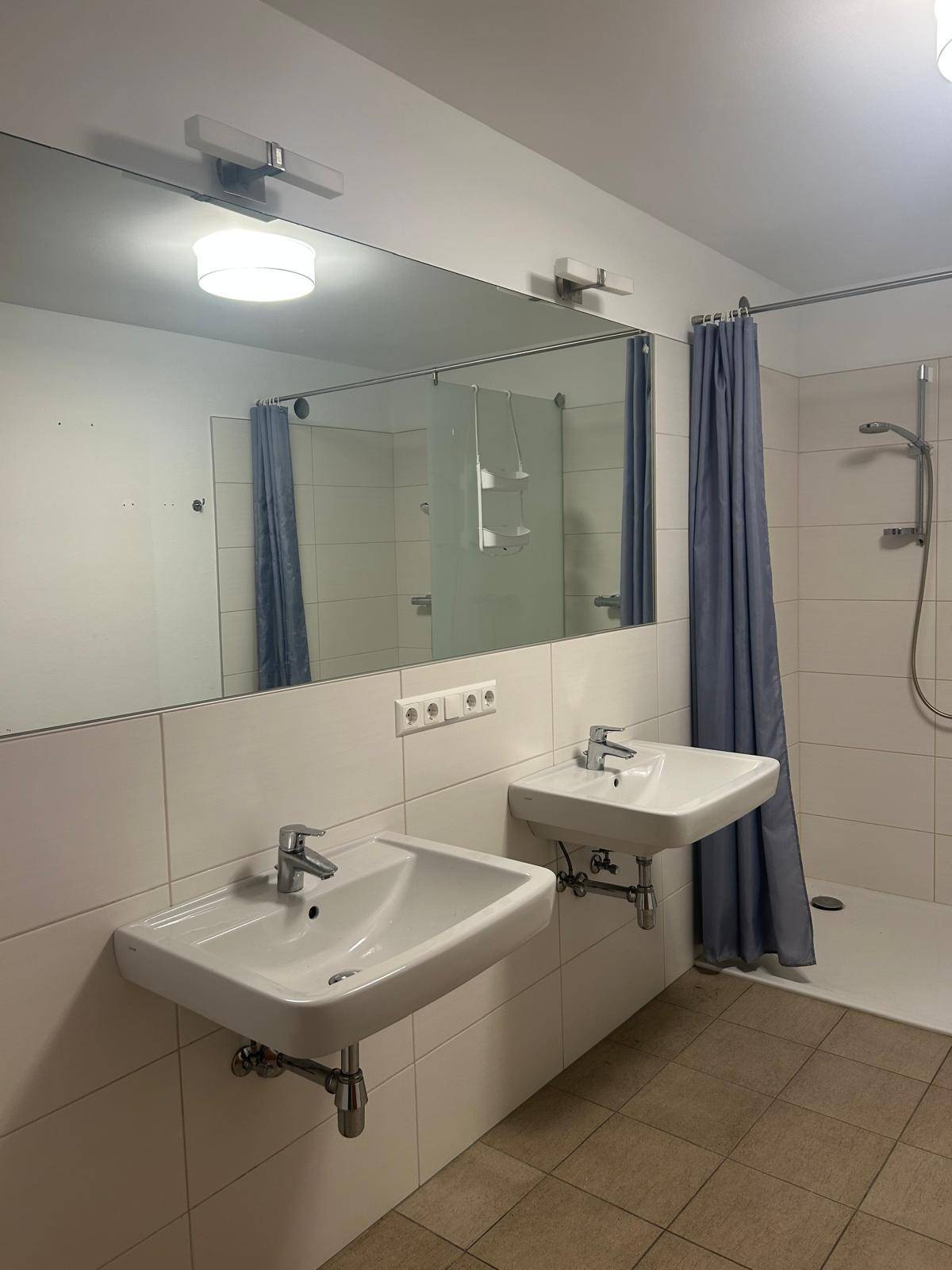 Badezimmer mit zwei Waschbecken, großem Spiegel und einer Duschkabine, ausgestattet mit hellen Fliesen.