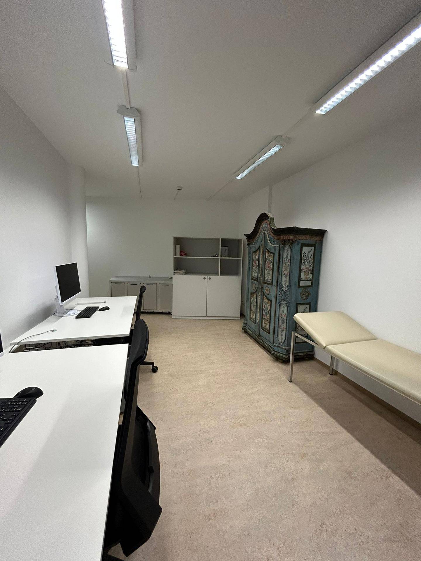 Büro mit Arbeitsplätzen, einem auffälligen antiken Schrank und einem Untersuchungsbett, hell und funktional.