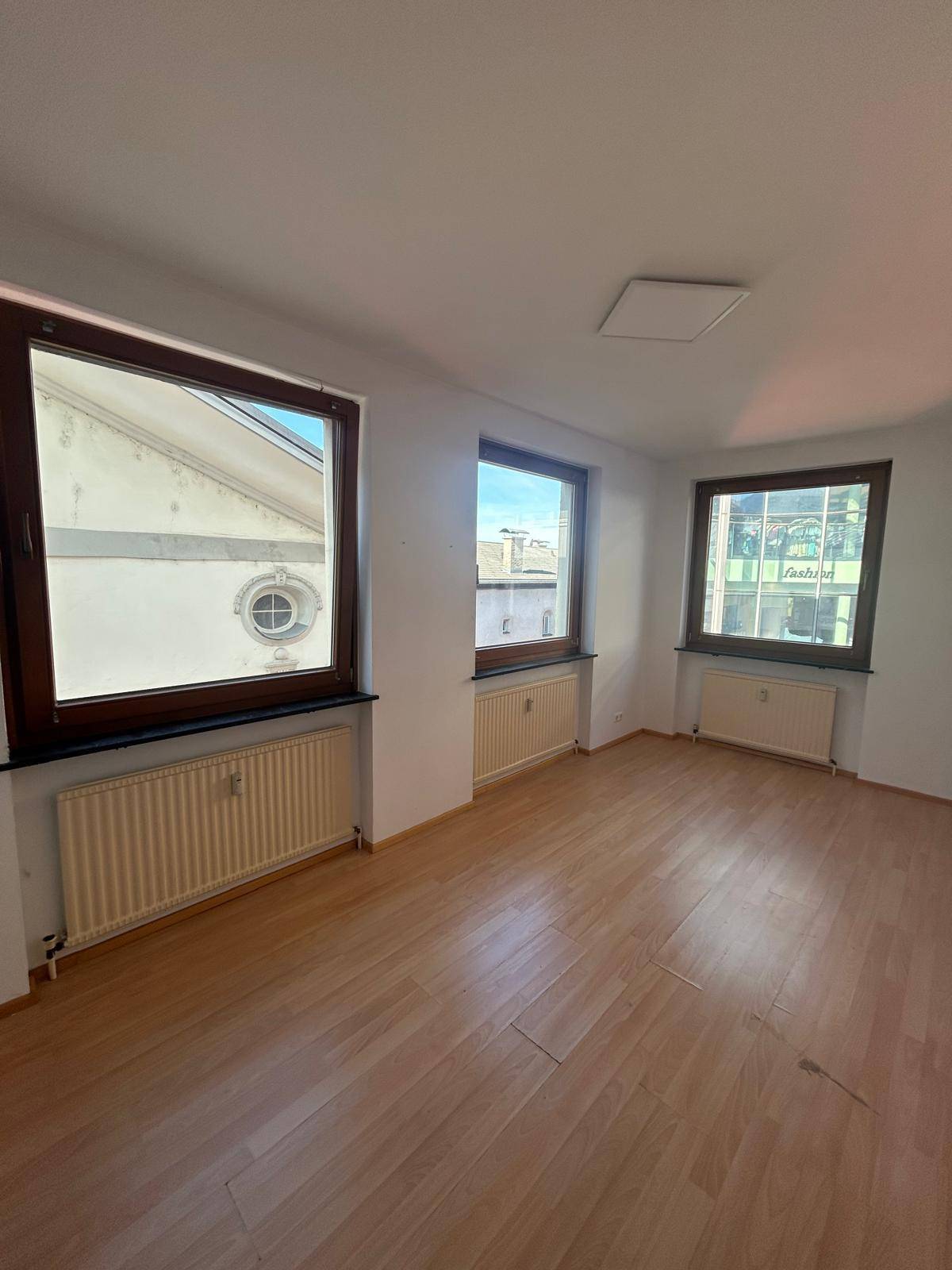 Heller Raum mit Laminatboden, drei Fenstern und Heizkörpern, bietet viel Tageslicht und Stadtblick.