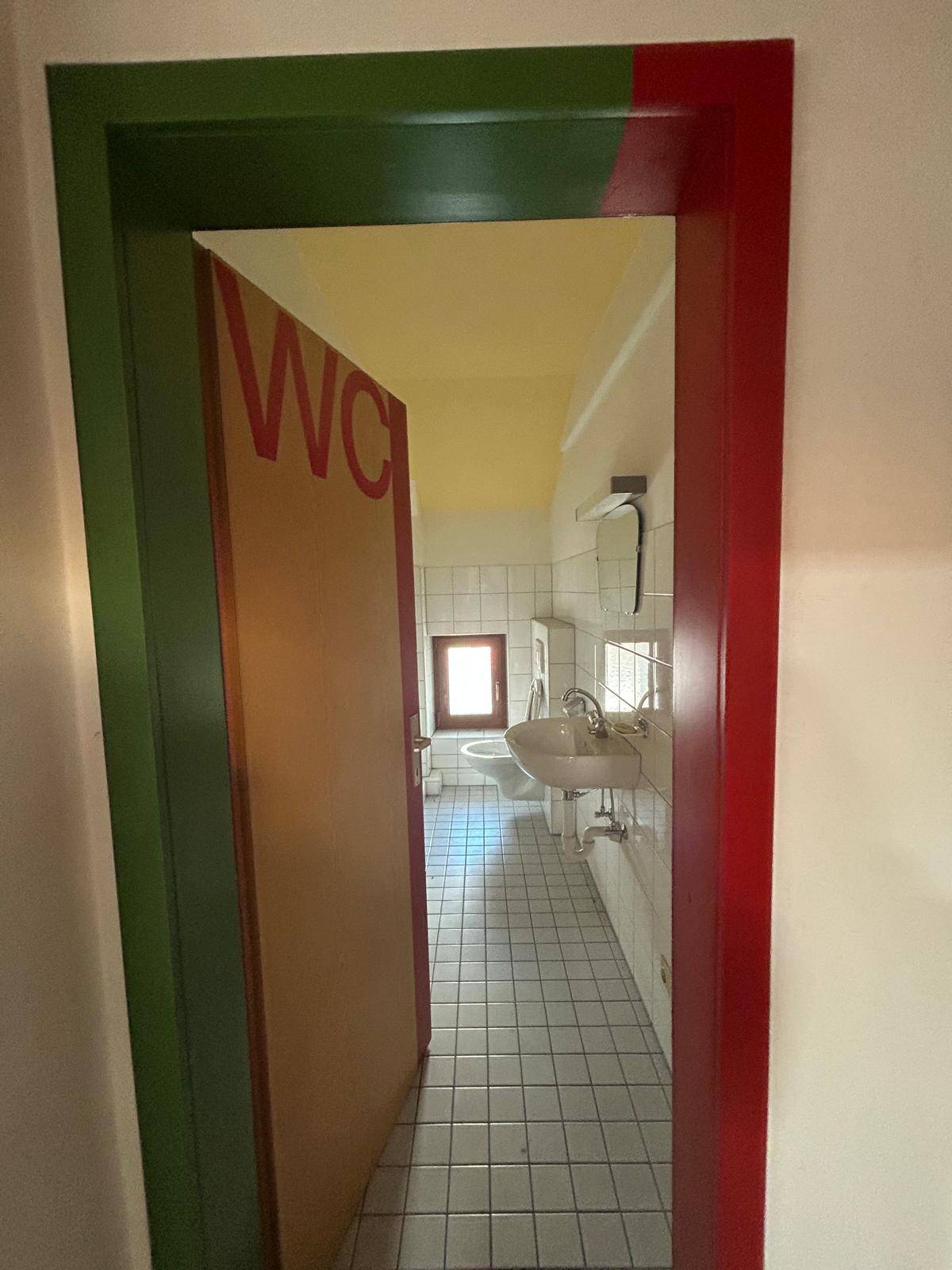 Einfache Toilette mit Fliesenboden, Waschbecken und Fenster, sauber und funktional.