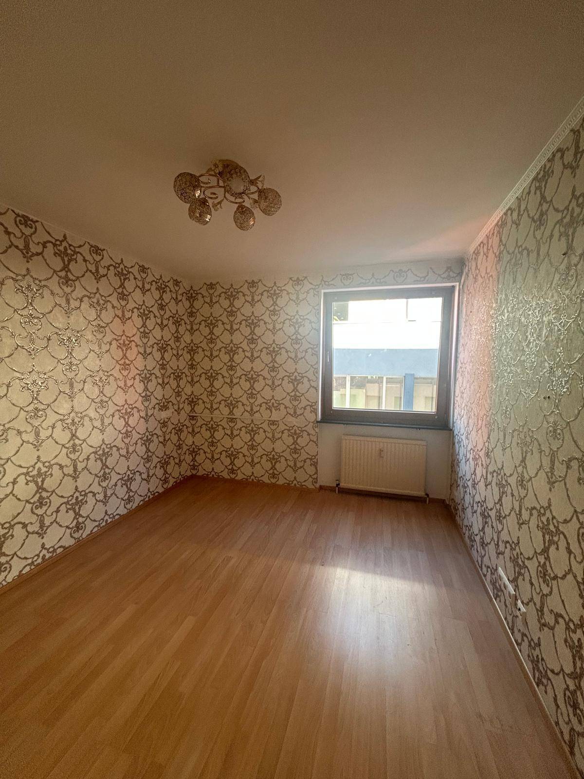 Zimmer mit auffälliger gemusterter Tapete, Laminatboden und einem Fenster, das Tageslicht hereinlässt.
