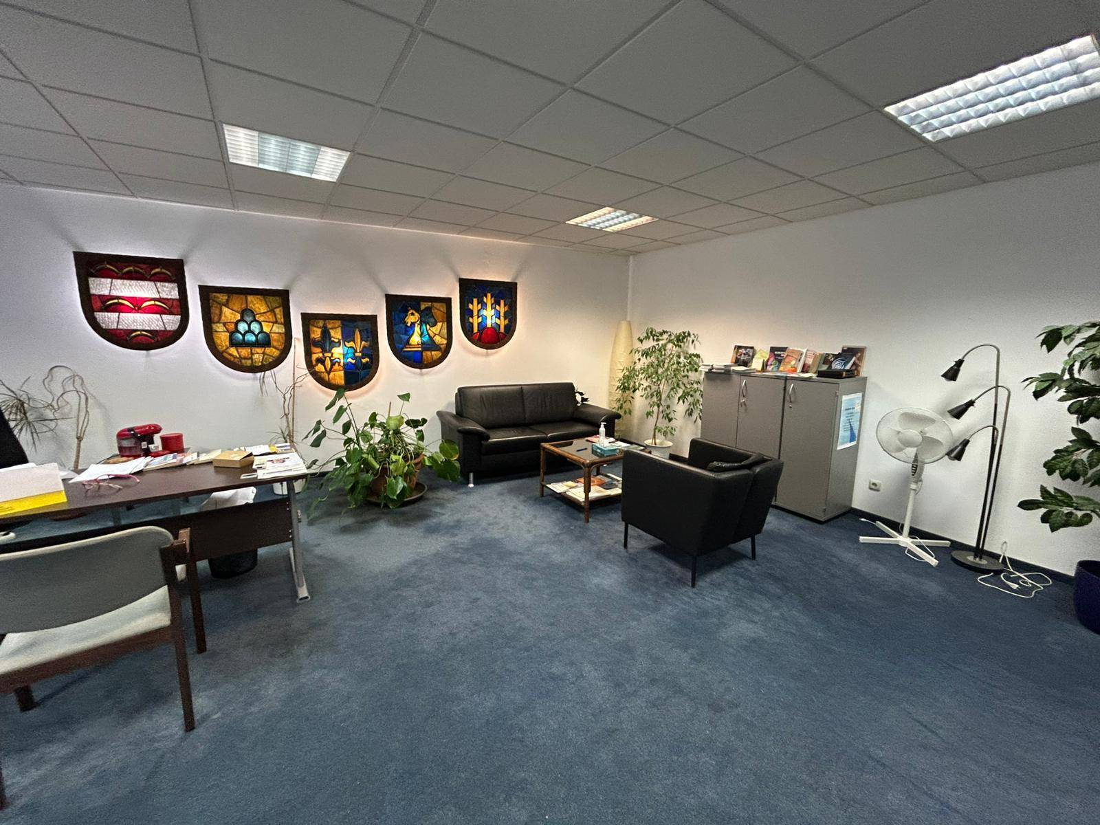 Großes Büro mit blauem Teppichboden, Schreibtisch, Ledersofa und dekorativen Glasfenstern an der Wand.