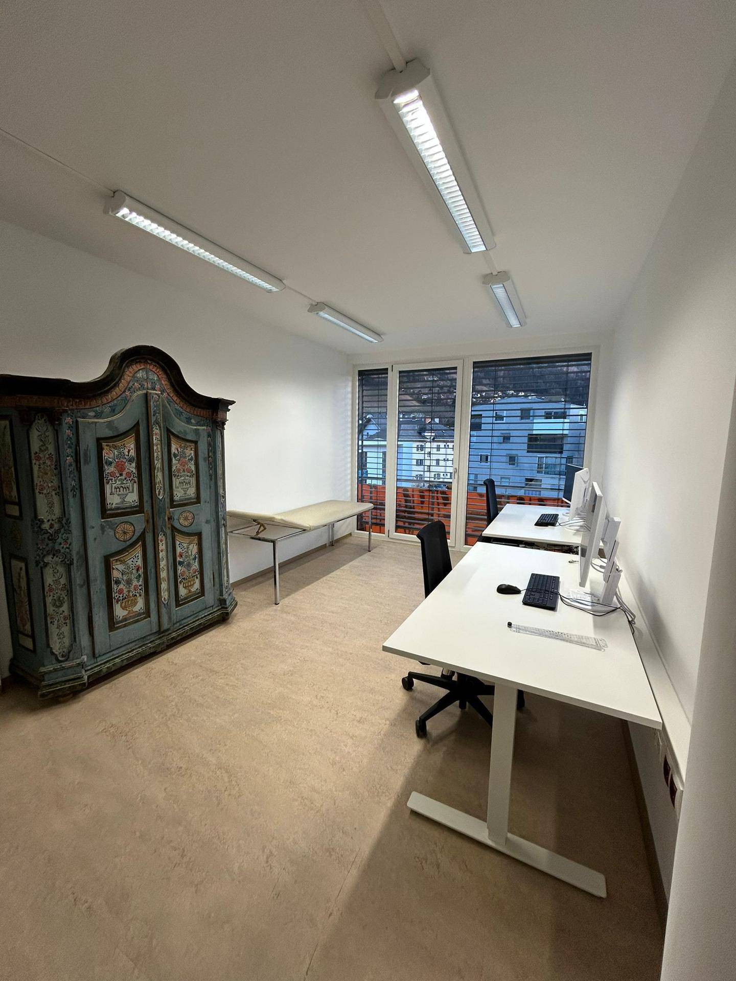 Geräumiges Büro mit mehreren Arbeitsplätzen, einem antiken Schrank und einem Untersuchungsbett, vielseitig nutzbar.