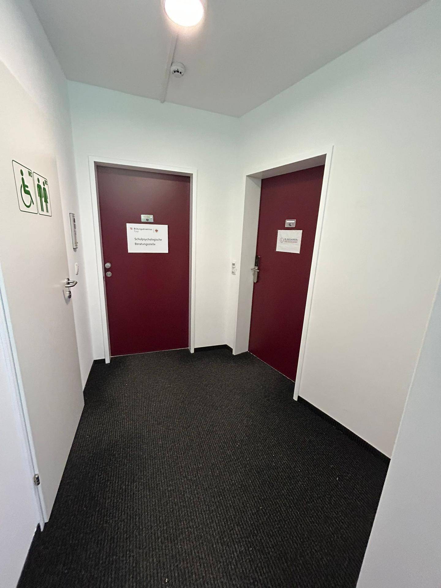 Schmaler Flur mit zwei markanten roten Türen und einem dunklen Teppichboden, der zu weiteren Räumen führt.