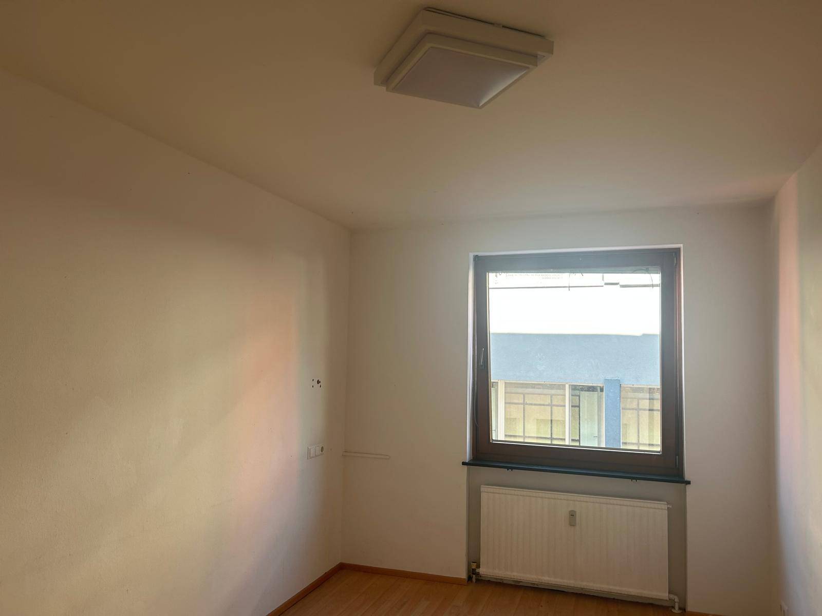 Heller Raum mit Laminatboden, weiß gestrichenen Wänden und einem Fenster mit Blick auf ein Gebäude.