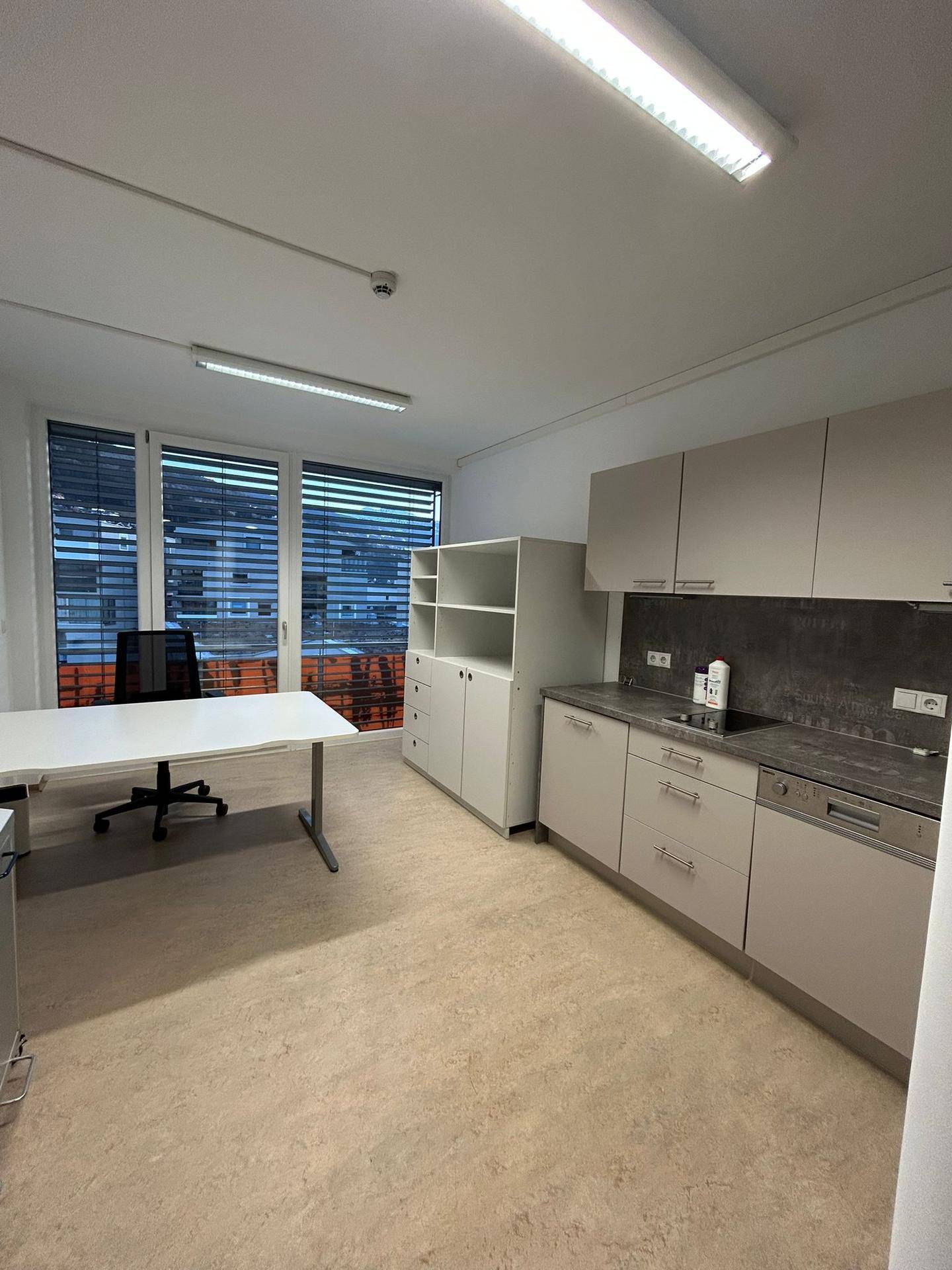 Modernes Büro mit integrierter Küchenzeile, großem Fenster und hellem Bodenbelag, ideal für effizientes Arbeiten.