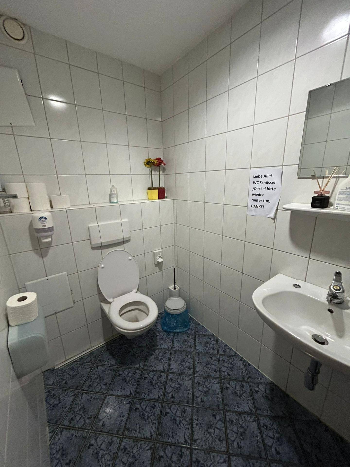 Gefliestes Badezimmer mit WC, Waschbecken und blauen Bodenfliesen, sowie einem Spiegel.