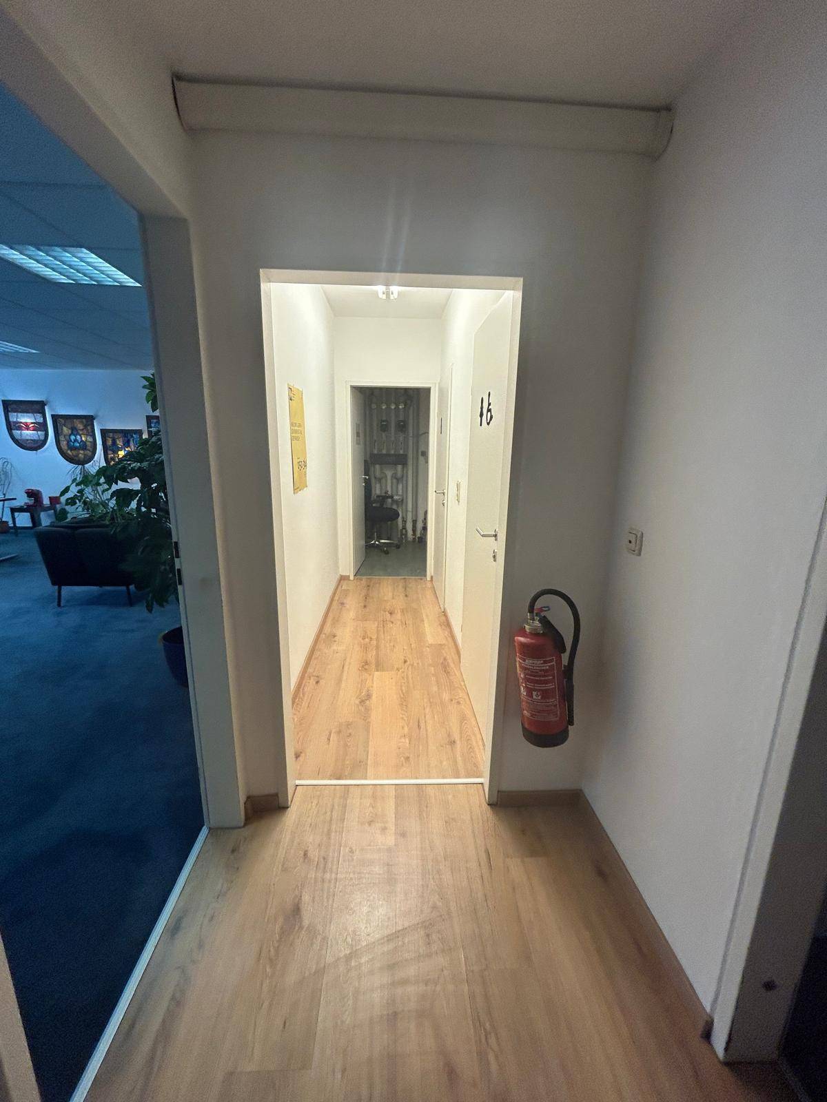 Langer Flur mit hellem Laminatboden, weißen Wänden und einem Feuerlöscher an der Wand.