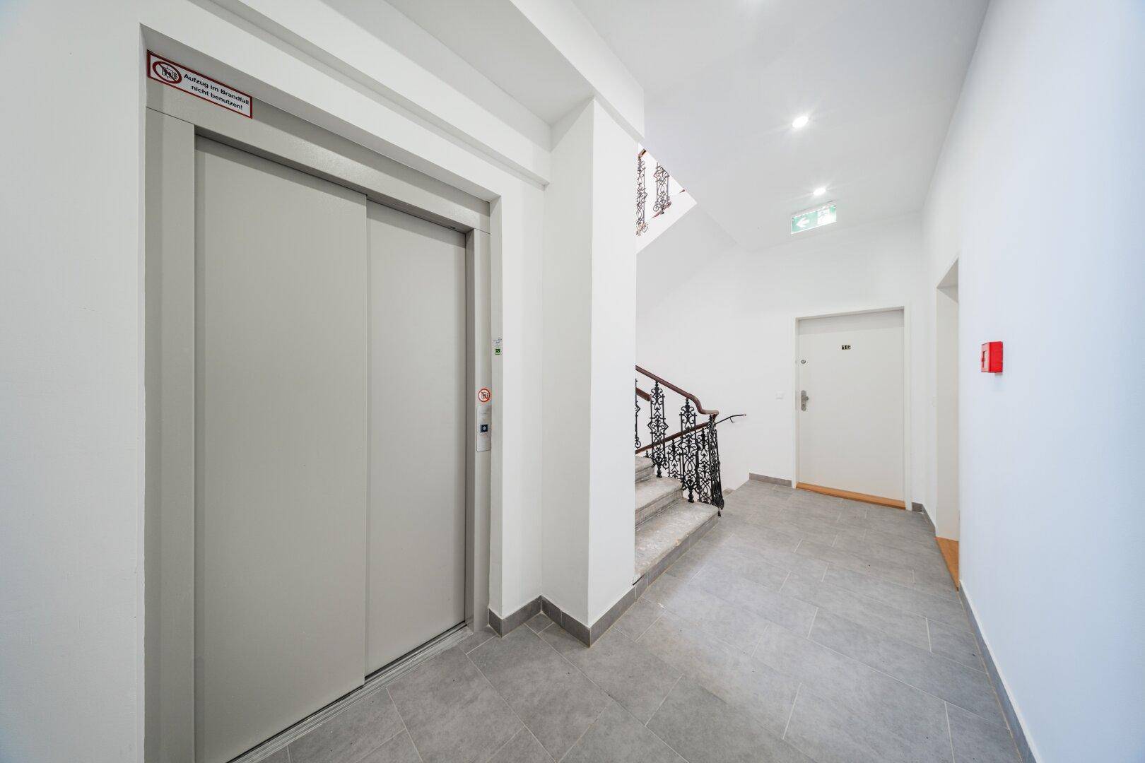 Moderner Aufzug in einem hellen Flur mit Zugang zum Treppenhaus und grauen Fliesen.
