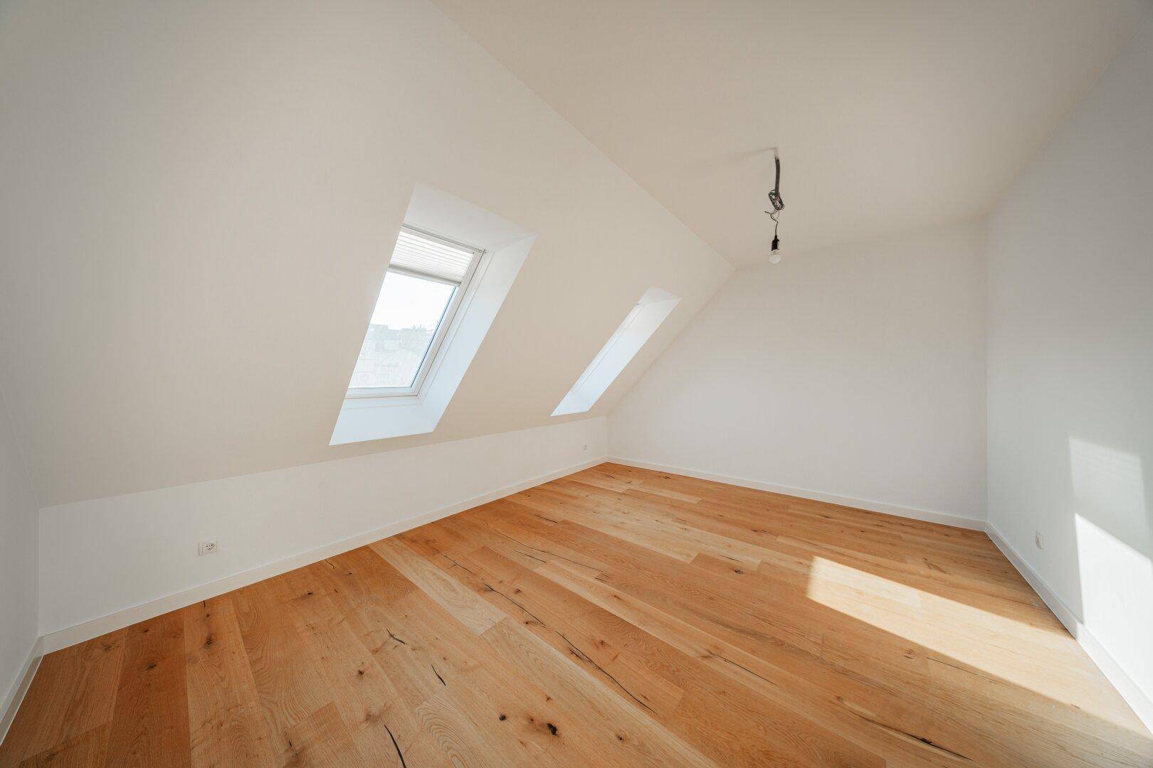 Leerer Dachgeschossraum mit Holzboden und zwei schrägen Dachfenstern, die für Helligkeit sorgen.