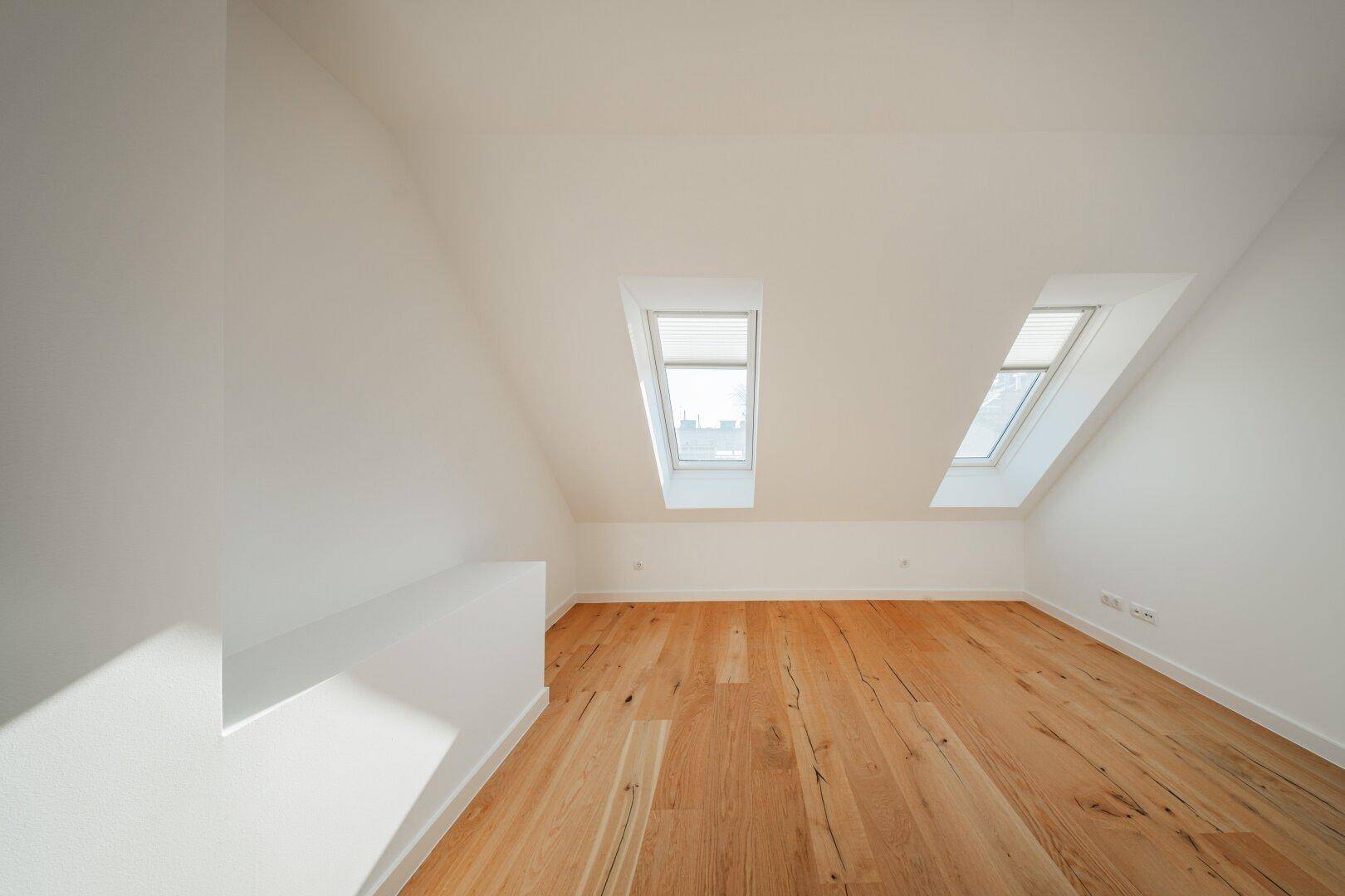 Heller Dachgeschossraum mit Holzboden und zwei Dachfenstern, die den Raum mit Licht füllen.