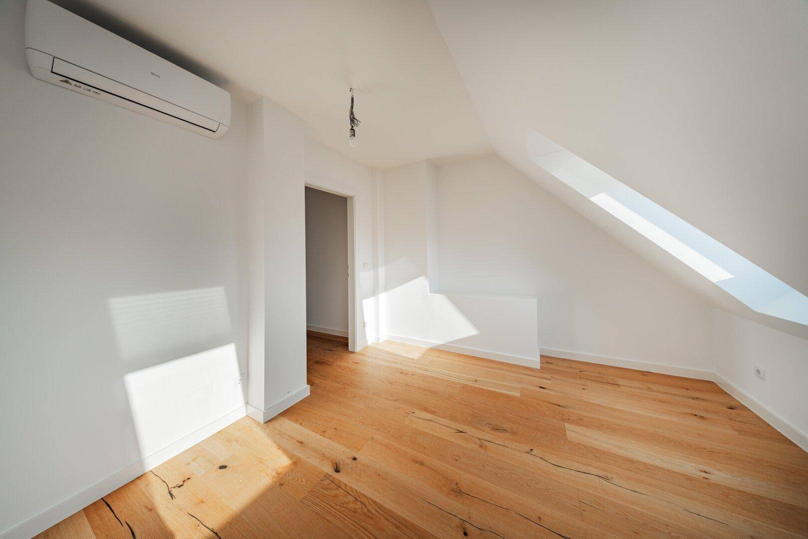 Leerer Raum im Dachgeschoss mit Holzboden, Klimaanlage und Zugang zu einem weiteren Bereich.