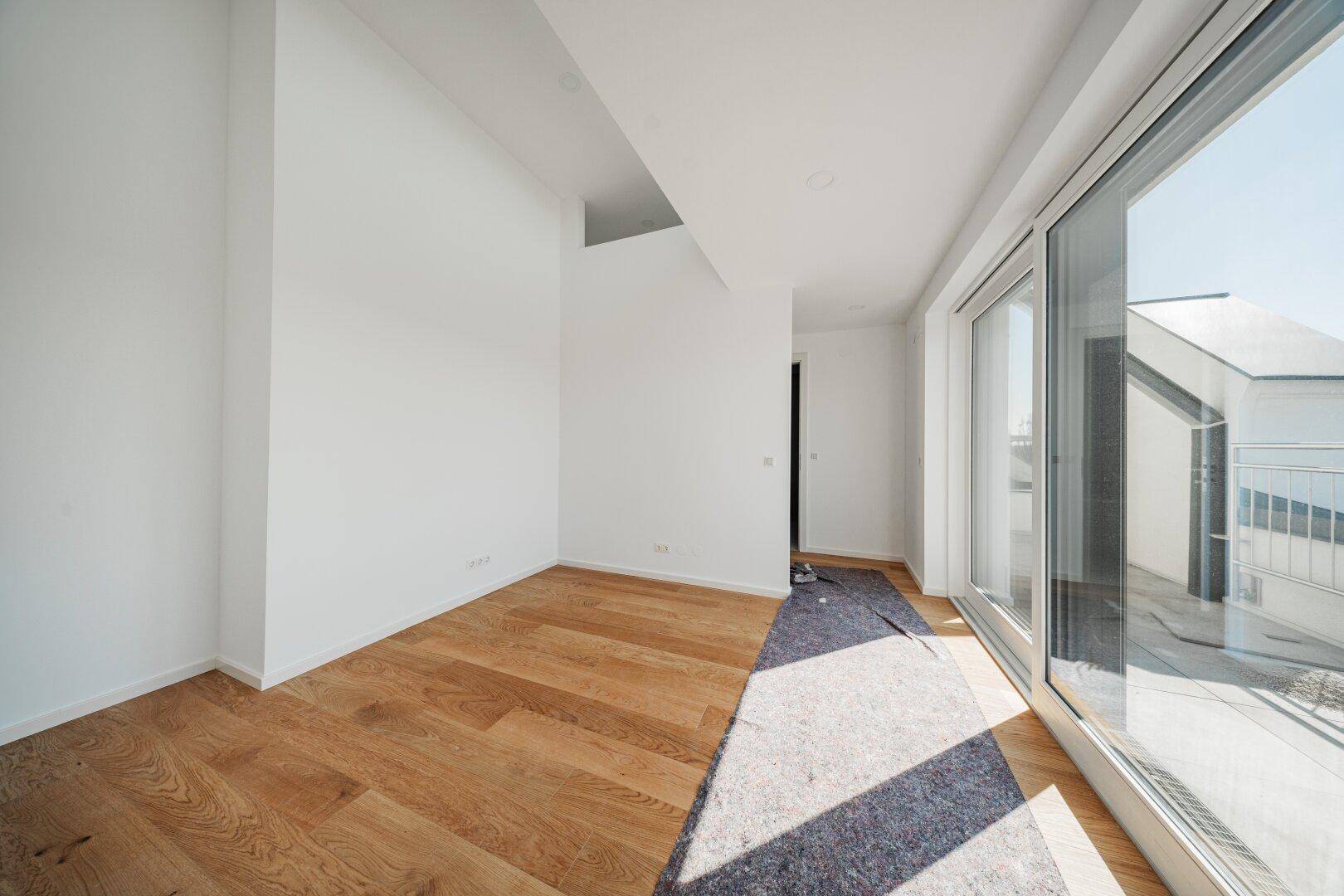 Heller, leerer Raum mit Holzboden und großen Fenstern, die den Blick nach draußen ermöglichen.