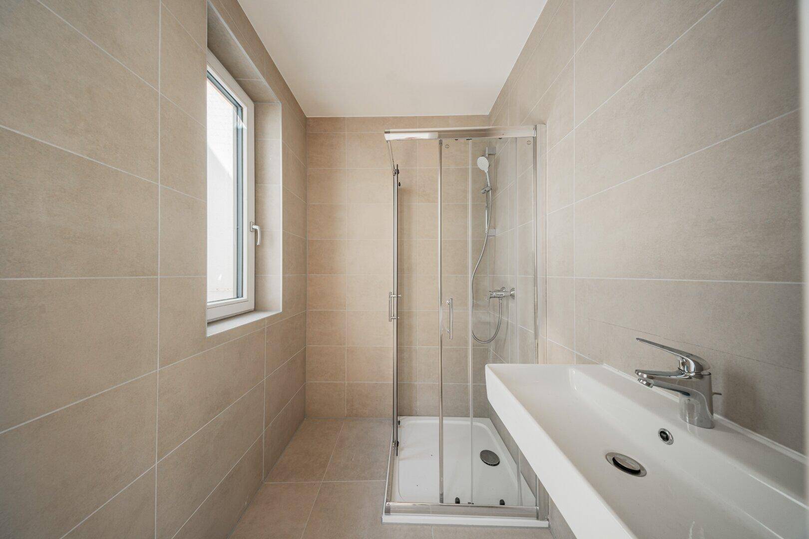 Modernes Badezimmer mit beigefarbenen Fliesen, Duschkabine und Waschtisch mit Fenster.