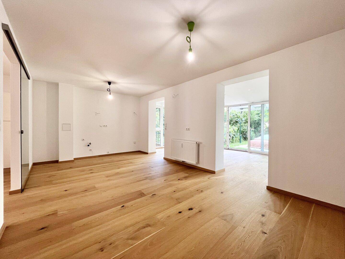 Offener Wohnbereich mit Holzboden, Heizkörper und Blick auf den grünen Außenbereich.