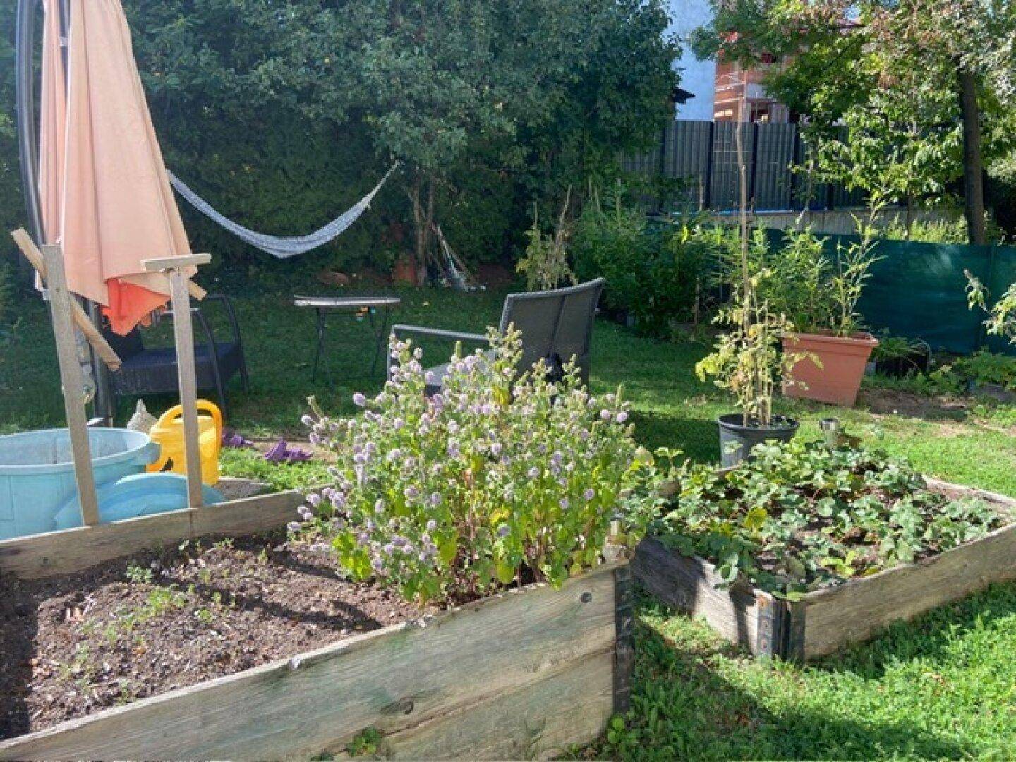 Grüner Garten mit Hochbeeten, Sonnenschirm und Hängematte, ideal für entspannte Stunden im Freien.