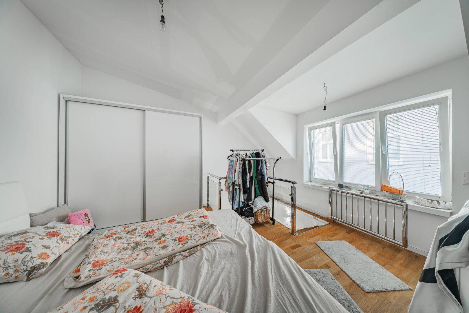 Helles Schlafzimmer mit Dachschräge, Holzboden und Fenstern mit Jalousien für viel Tageslicht.