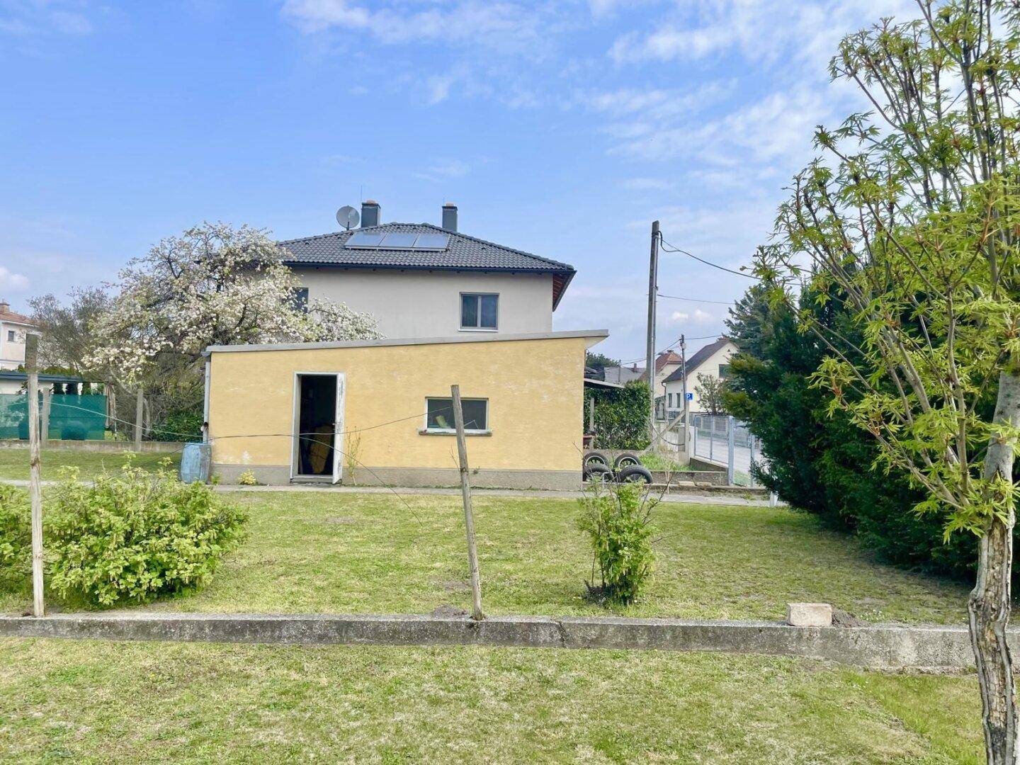 Gelber Gartenschuppen im Grünen mit einem Nachbarhaus im Hintergrund, das Solarpaneele besitzt.