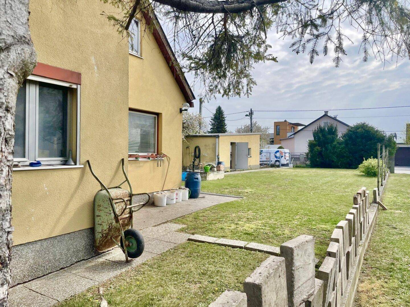 Seitenansicht des Hauses mit Garten, Zaun und einer Schubkarre im Vordergrund.